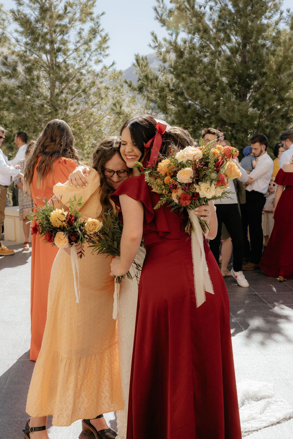 Bridesmaids in warm-toned dresses share joyful hugs while holding colorful wedding bouquets.