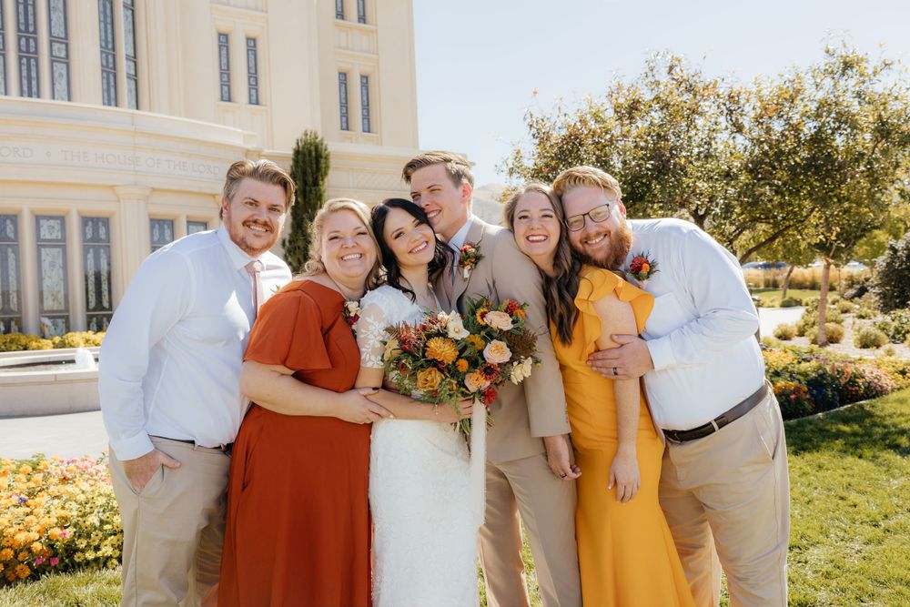 Small wedding party group smiling together outdoors in front of a white temple on a sunny day.