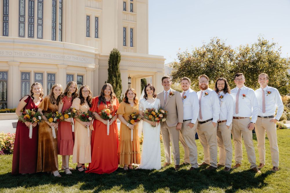 Large wedding party posing together outside a white temple with bridesmaids in fall-colored dresses.