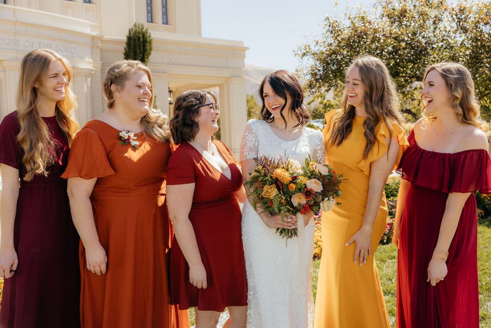 Bride in white lace gown holding bouquet with bridesmaids in red, orange, and yellow dresses.