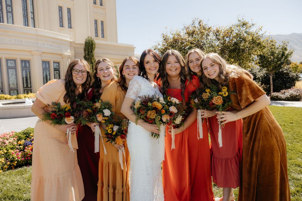 Bride surrounded by bridesmaids in mismatched fall-toned dresses holding floral bouquets outside.