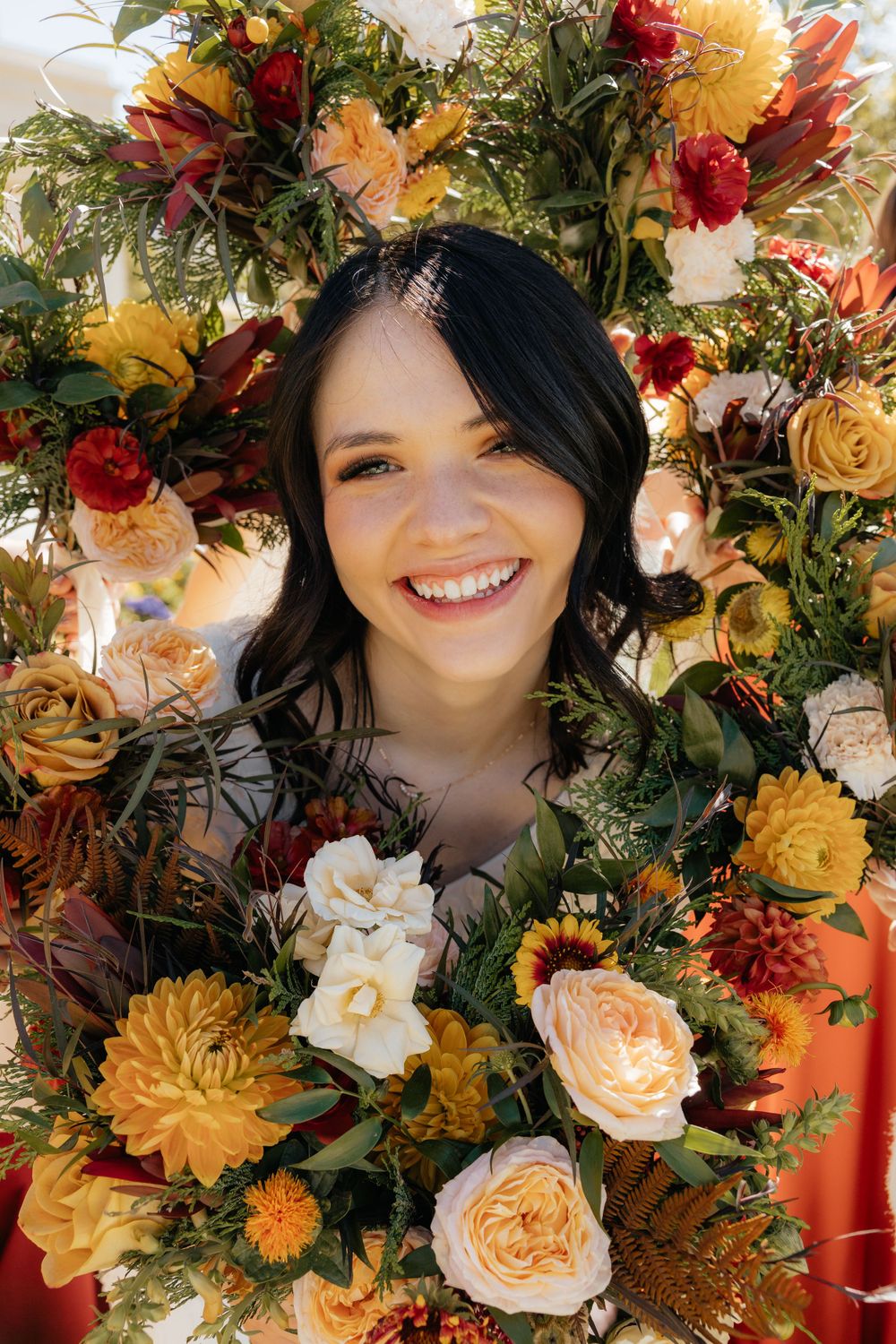 Smiling bride framed by vibrant fall floral arrangement with roses and dahlias in warm tones.