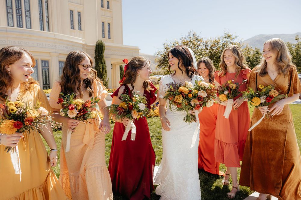 Bride and bridesmaids in autumn-colored dresses laughing together on a sunny temple lawn.