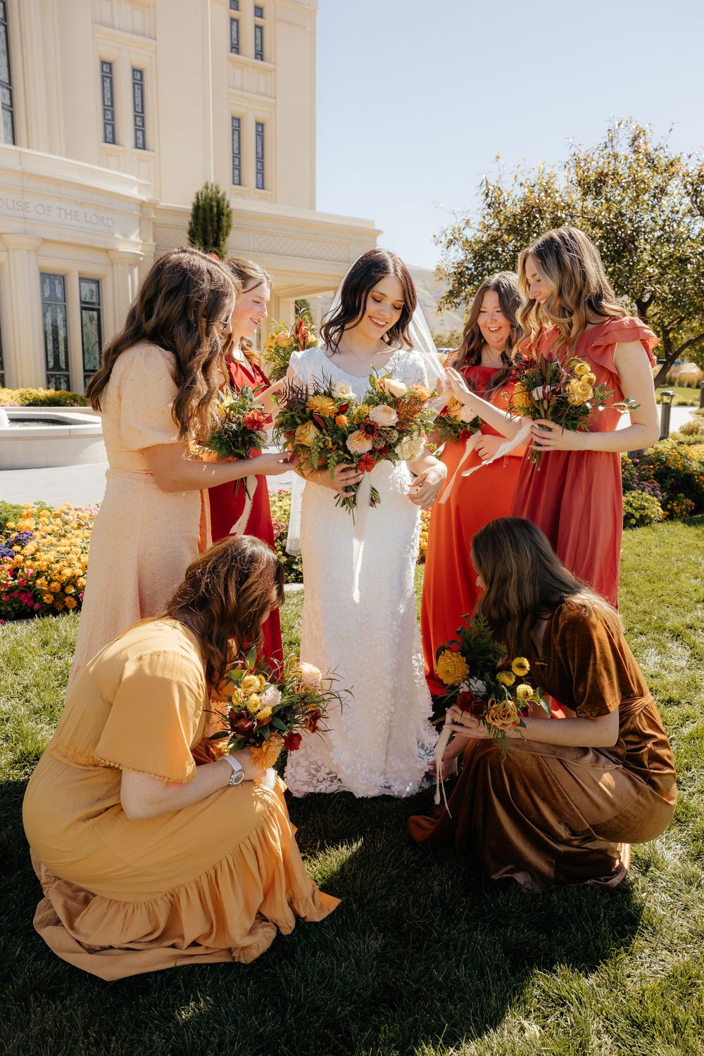Bride surrounded by bridesmaids in autumn-toned dresses admiring colorful fall bouquets on a sunny temple lawn.