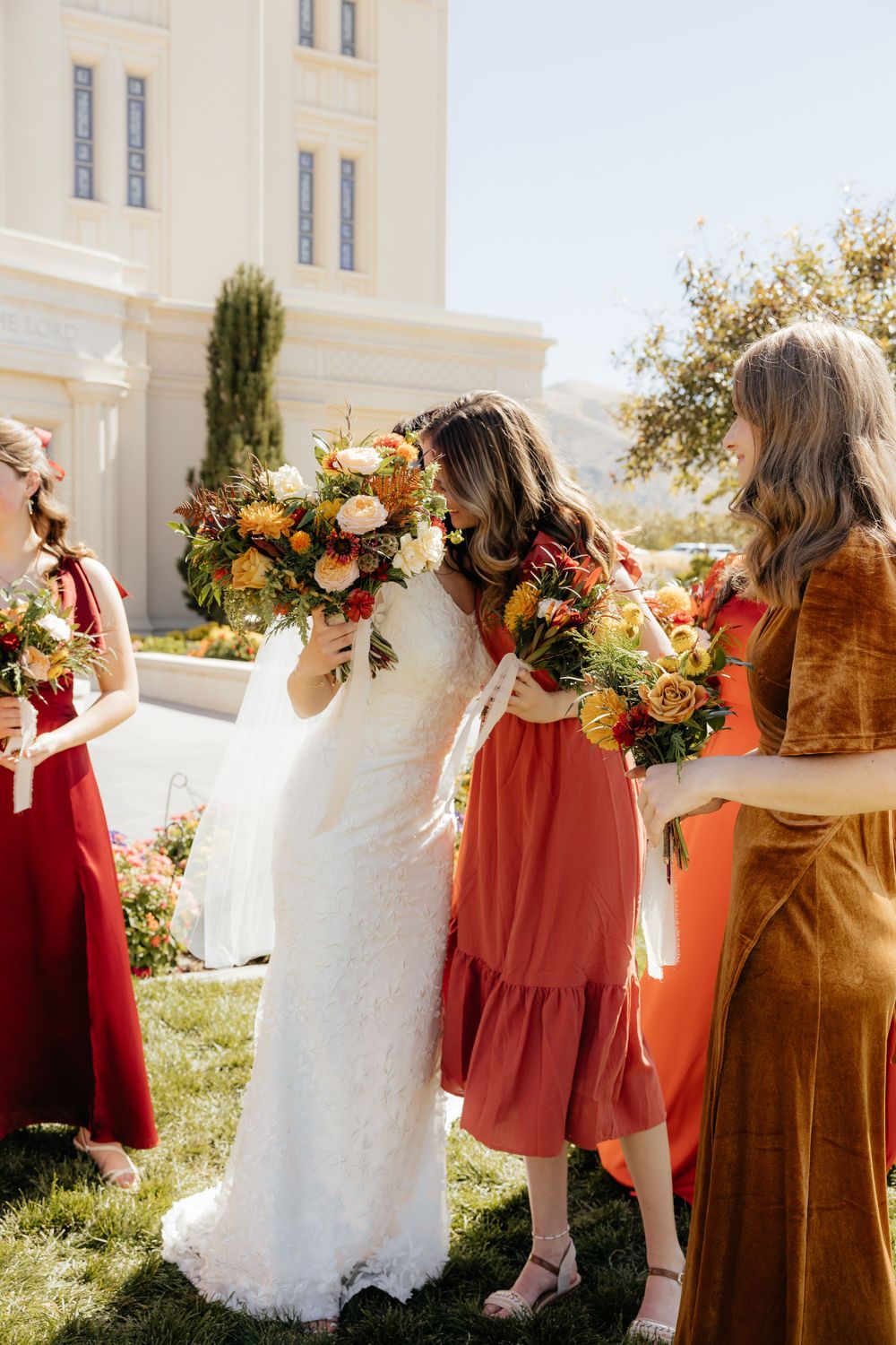 Bride in white lace gown smelling her bouquet surrounded by bridesmaids in fall dresses.