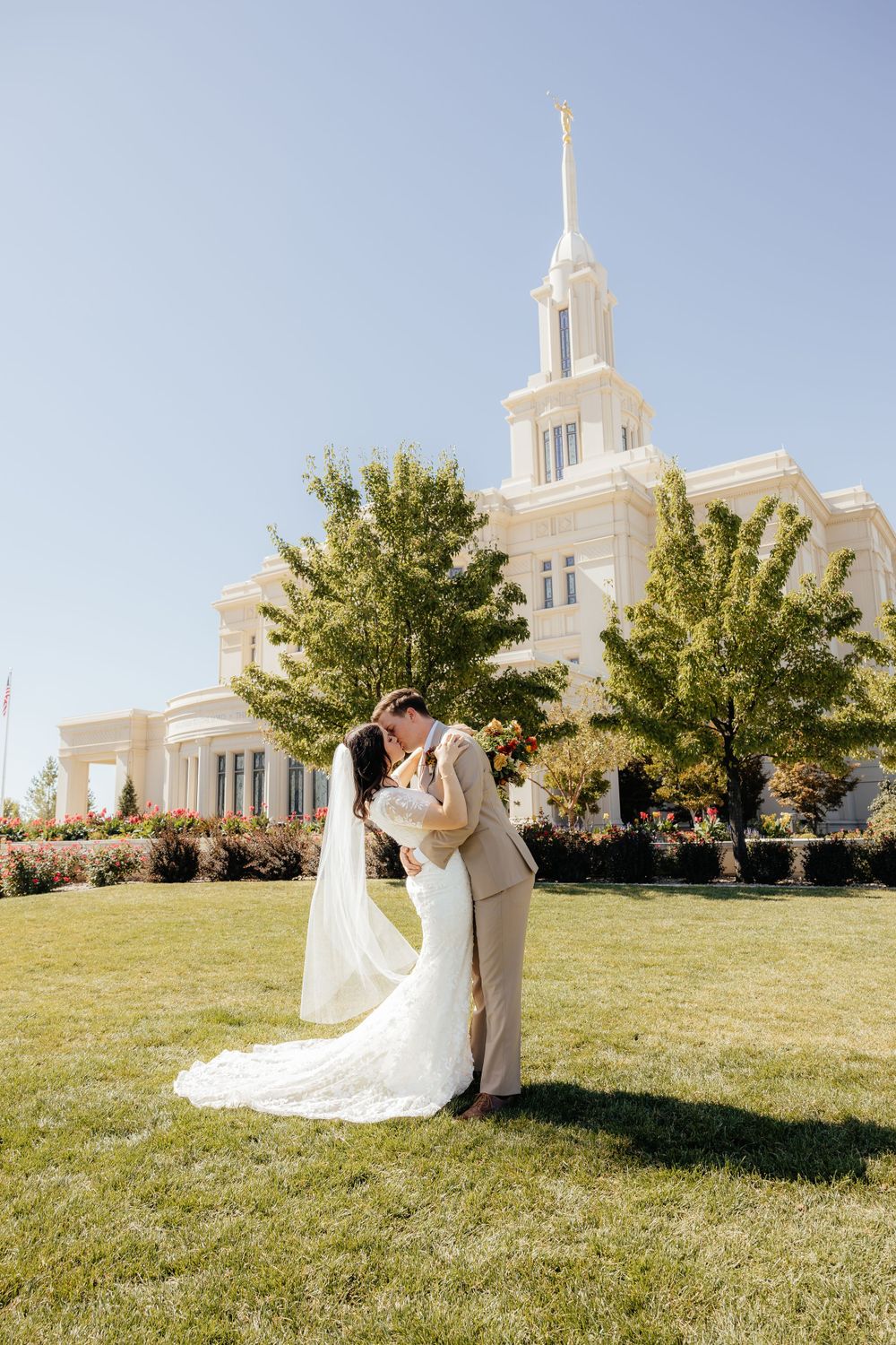 Bride and groom sharing a romantic kiss on the lawn in front of a white LDS temple on a sunny day.