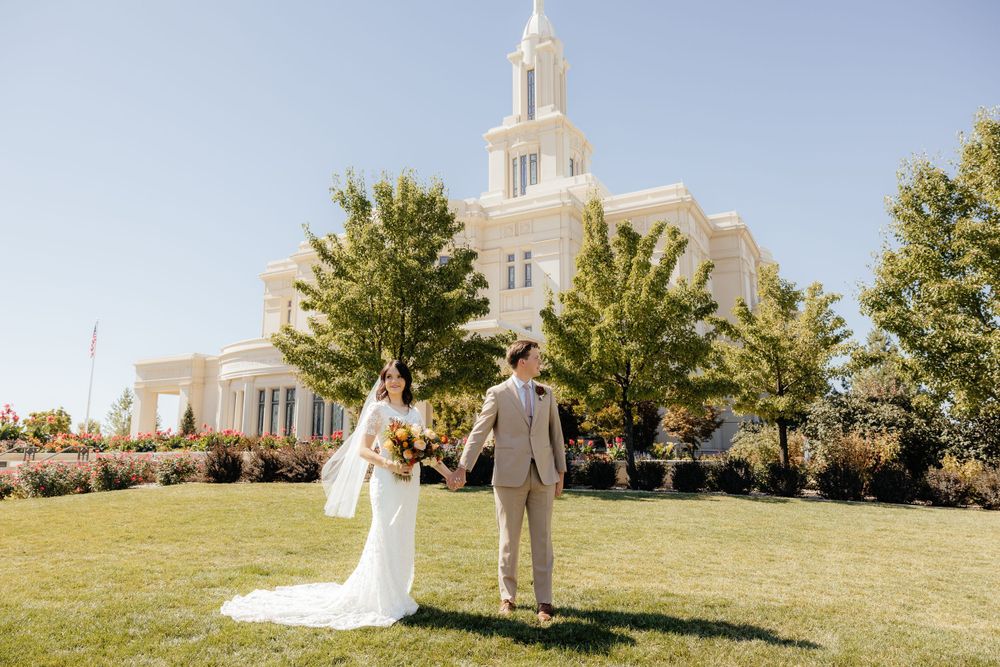 Bride and groom holding hands while posing on green grass in front of a grand white LDS temple.