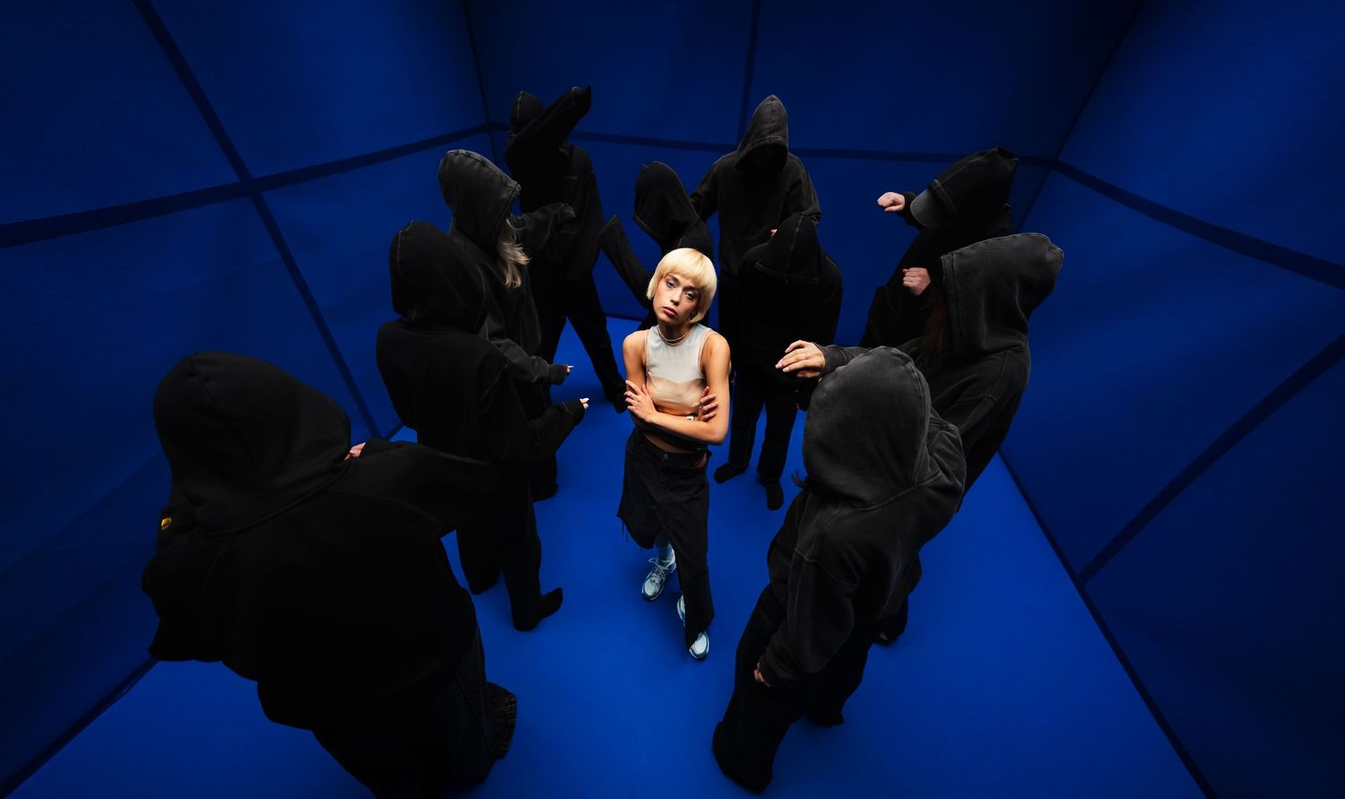 Music video or editorial photo of a woman in a white top surrounded by figures in black hooded outfits in a vivid blue room