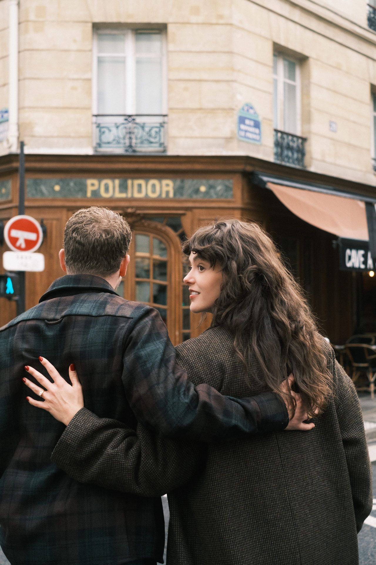 A couple in dark coats embraces as they walk along a Parisian street with vintage storefronts visible.
