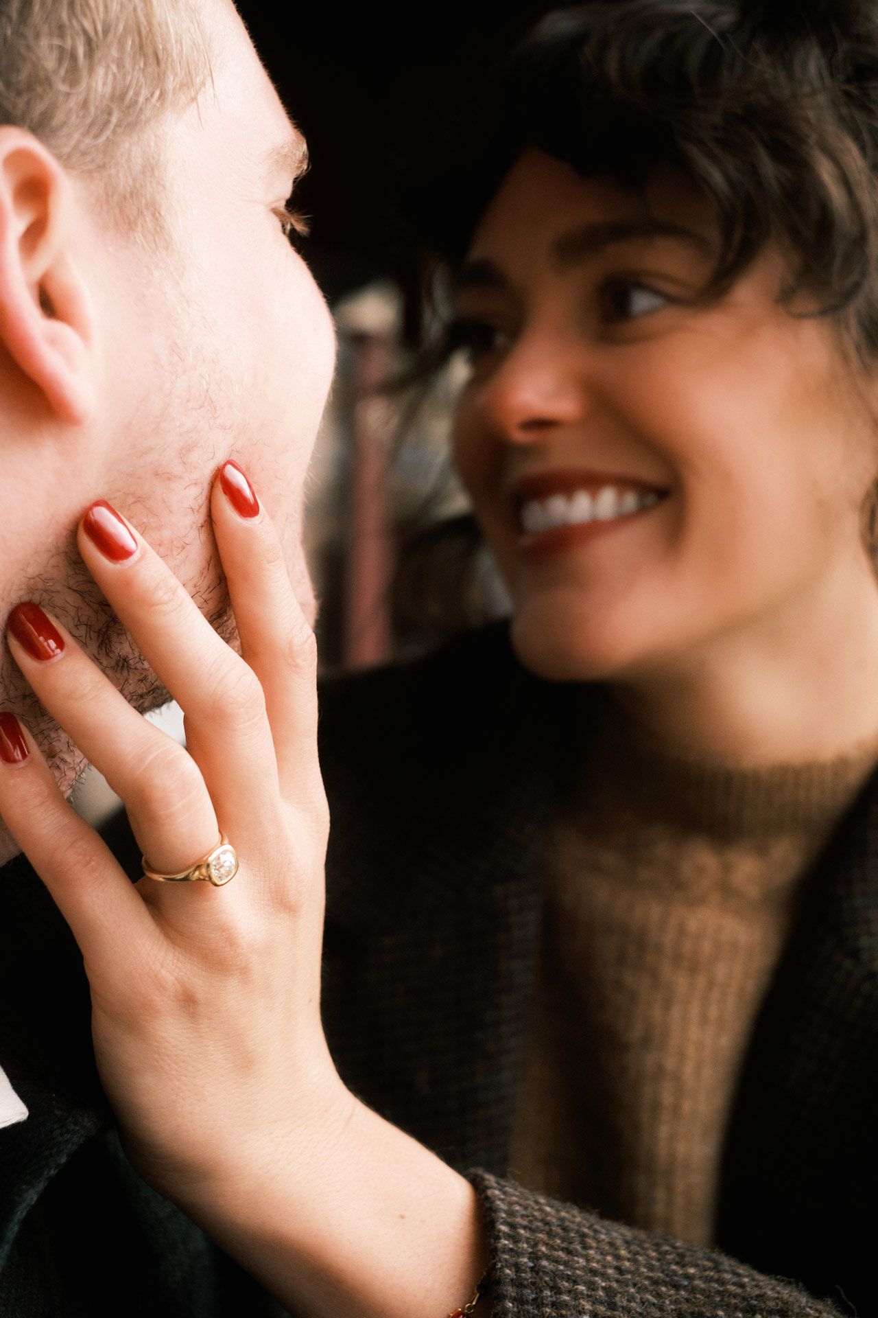 Close-up intimate moment showing person in sweater caressing another's face with red painted nails and engagement ring.