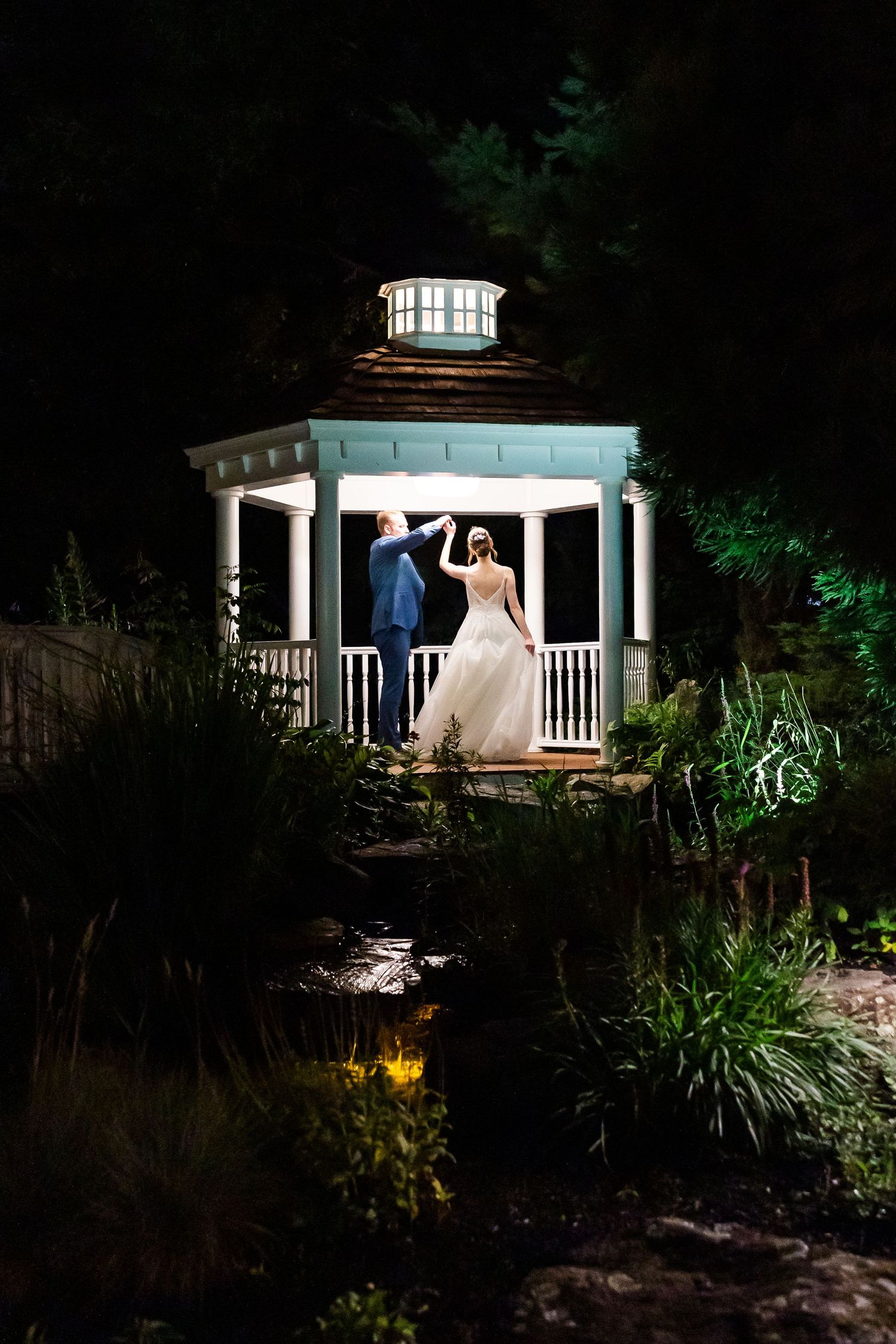 A white gazebo illuminated at night in a garden setting with landscape lighting creating a romantic atmosphere.