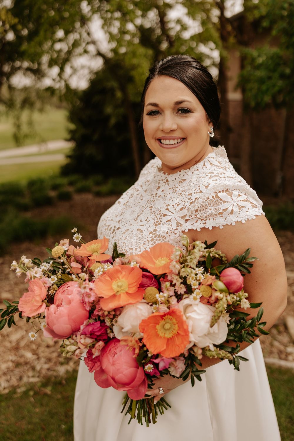 Elegant bride in white lace dress holding vibrant coral and pink peony bouquet in outdoor garden setting.