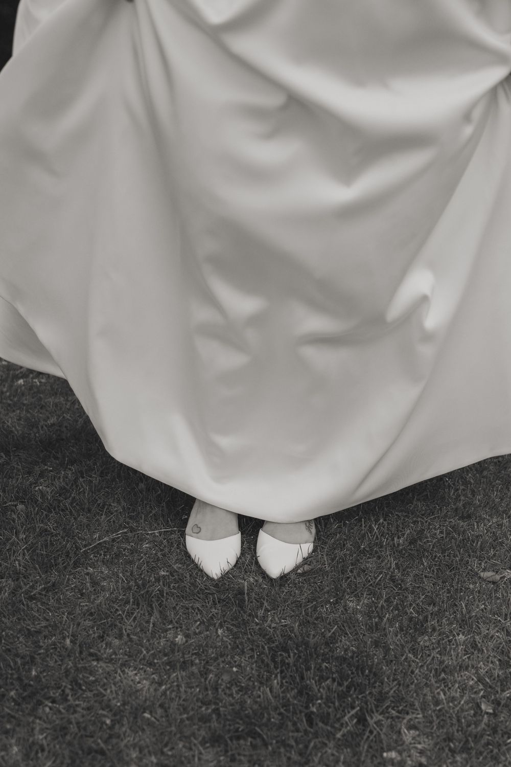 A close-up black and white photo of white bridal shoes peeking out from beneath a flowing wedding dress.
