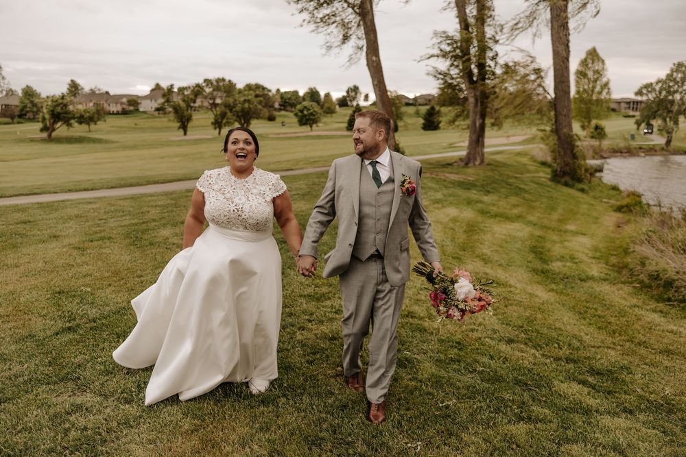 A joyful couple walks hand in hand across a lush golf course on their wedding day, wearing traditional white gown and grey suit.