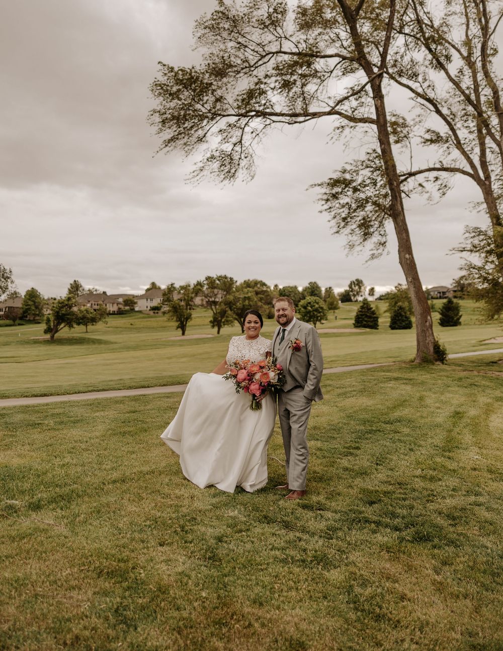 A wedding couple poses on a lush golf course beneath a large tree on an overcast day.