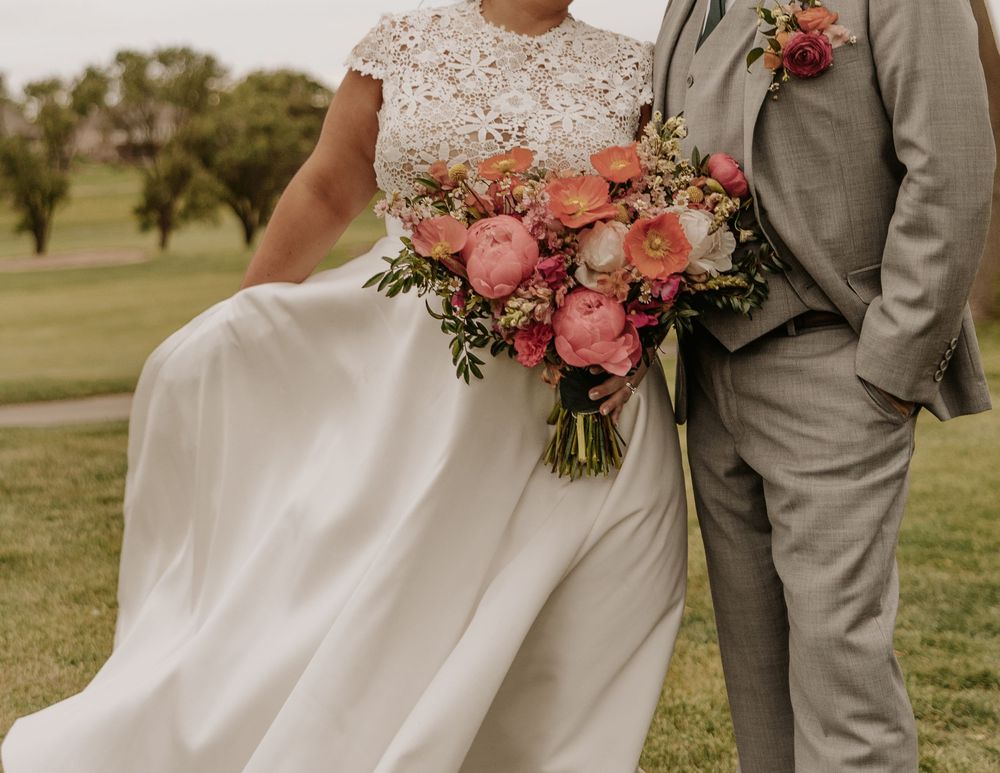 A wedding couple poses together on a grassy field holding a bouquet of pink and coral flowers with greenery.