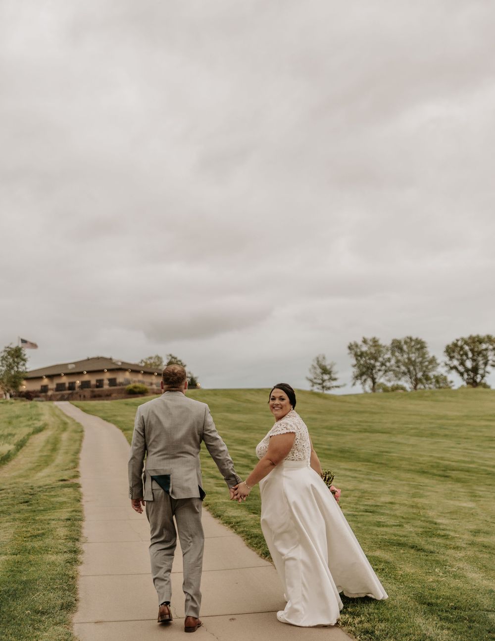 A bride and groom walk hand in hand down a winding path on a rolling green golf course under cloudy skies.