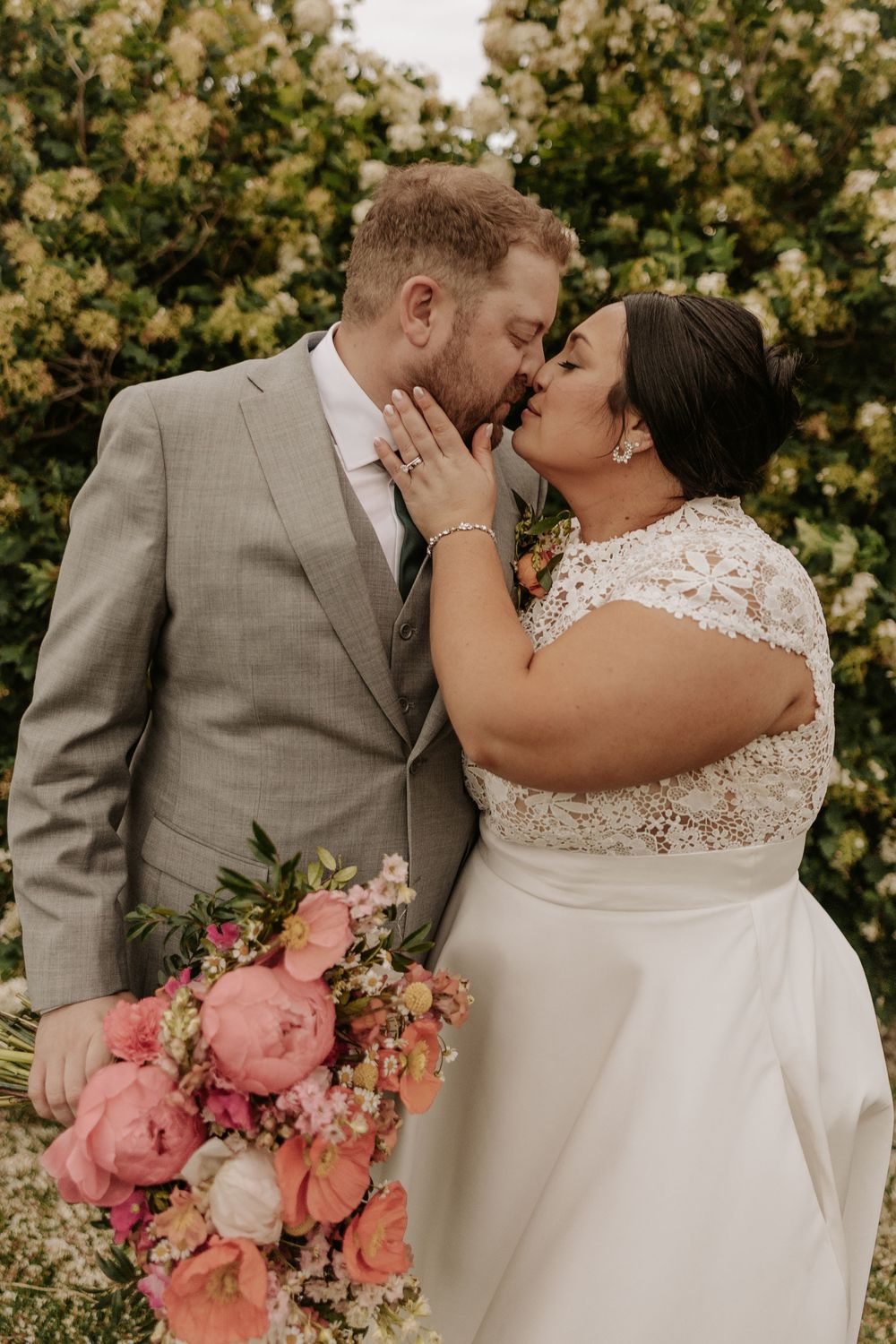 A romantic kiss between newlyweds in formal attire with a beautiful coral and pink wedding bouquet against flowering bushes.