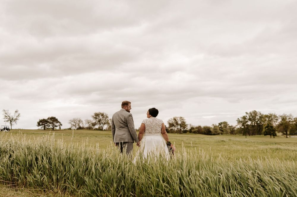 Two people walking hand in hand through a tall golden grass field at sunset against a cloudy sky.