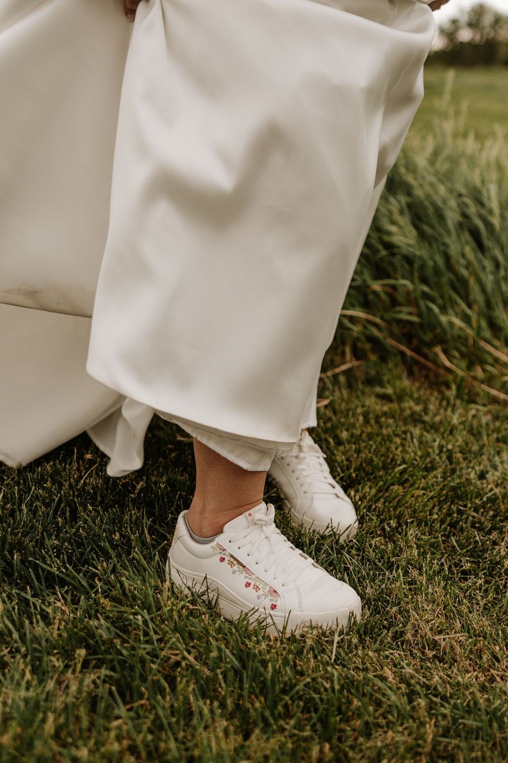 White sneakers worn with flowing white pants stand out against lush green grass in an outdoor setting.
