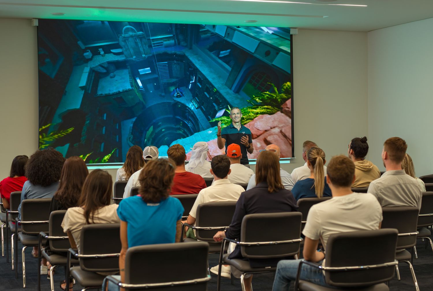 Audience members seated in rows of chairs watch a presentation in front of a blue painted mural wall.
