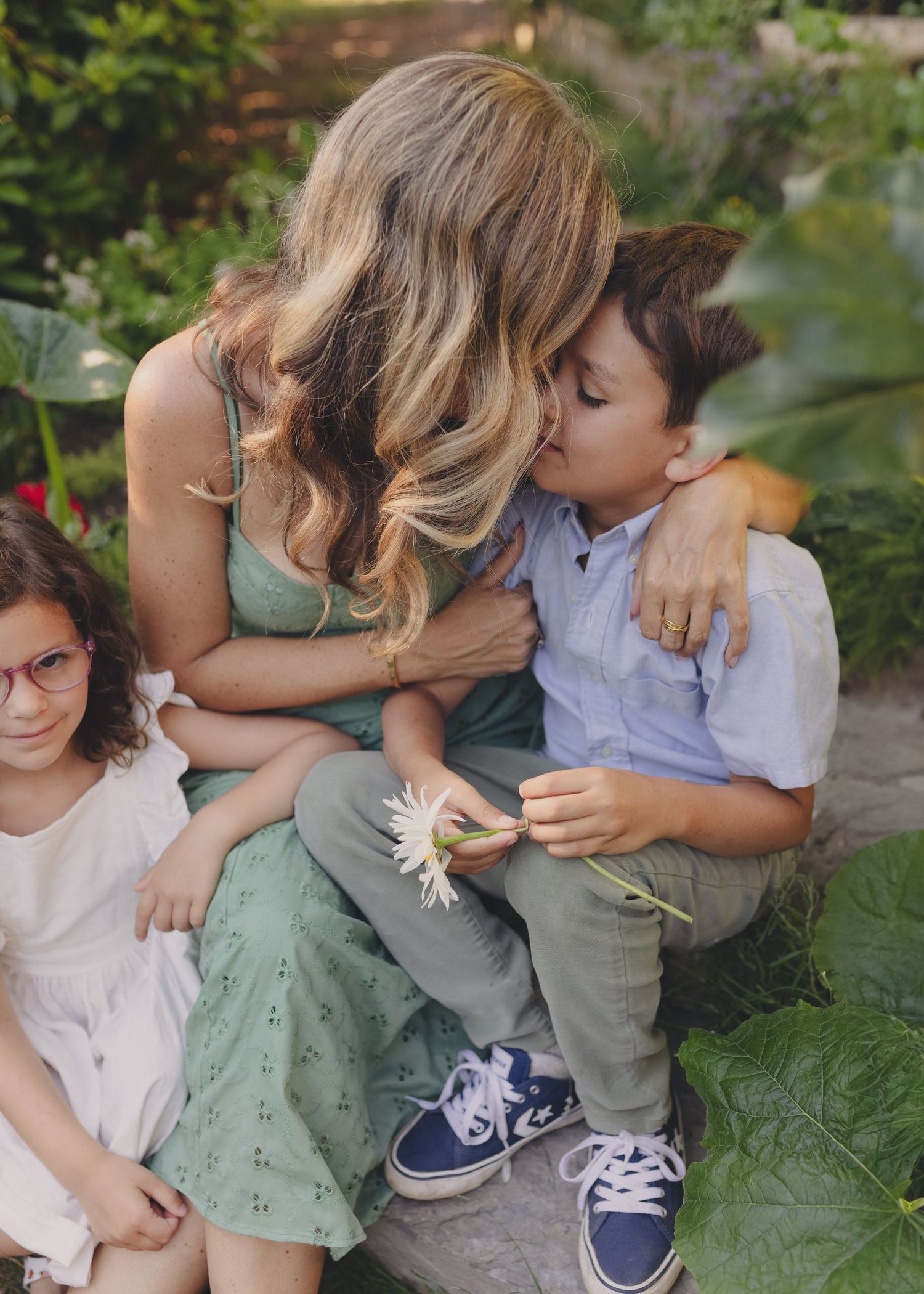 garden mini session Candice and kids sit on a blanket under the trees that glow with summer sunshine. mom kisses her boy