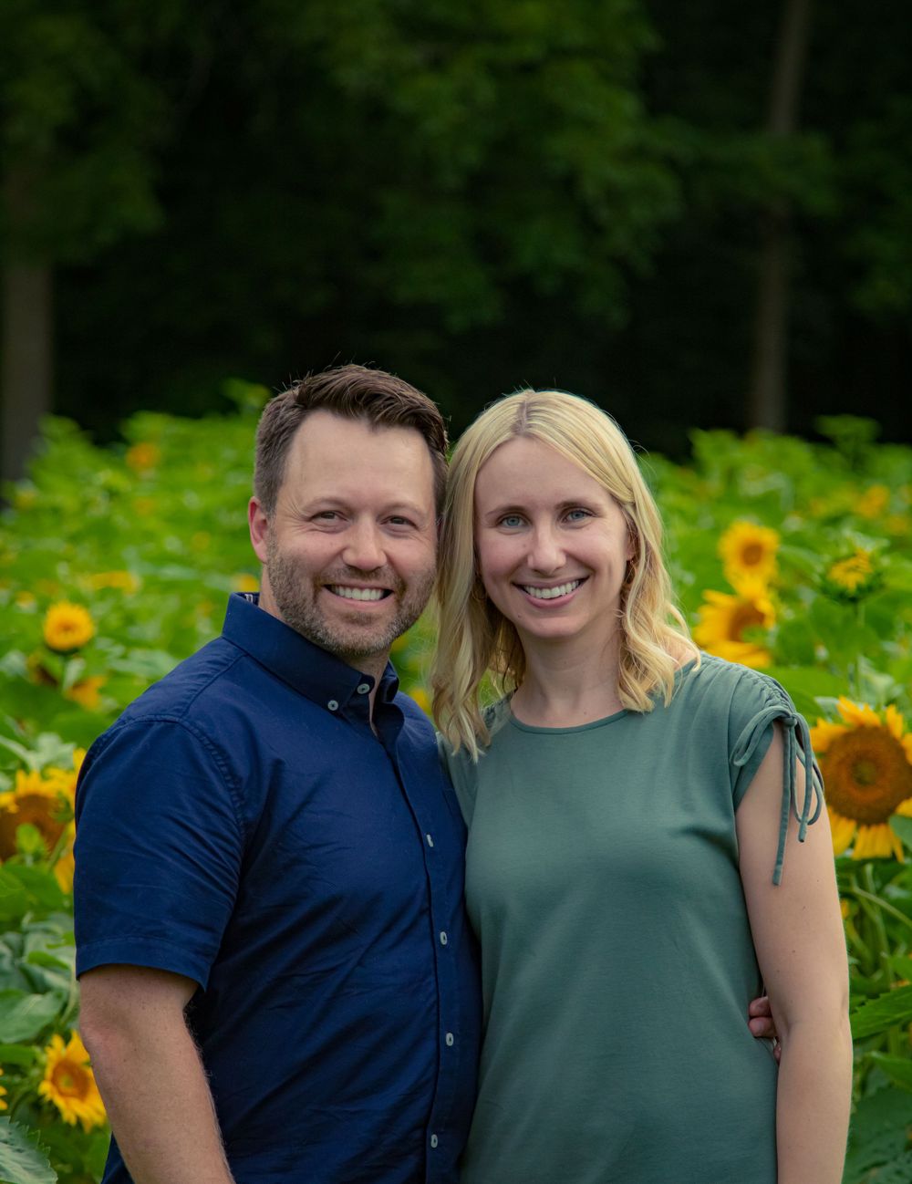 A couple poses together in front of a vibrant sunflower field on a sunny summer day.
