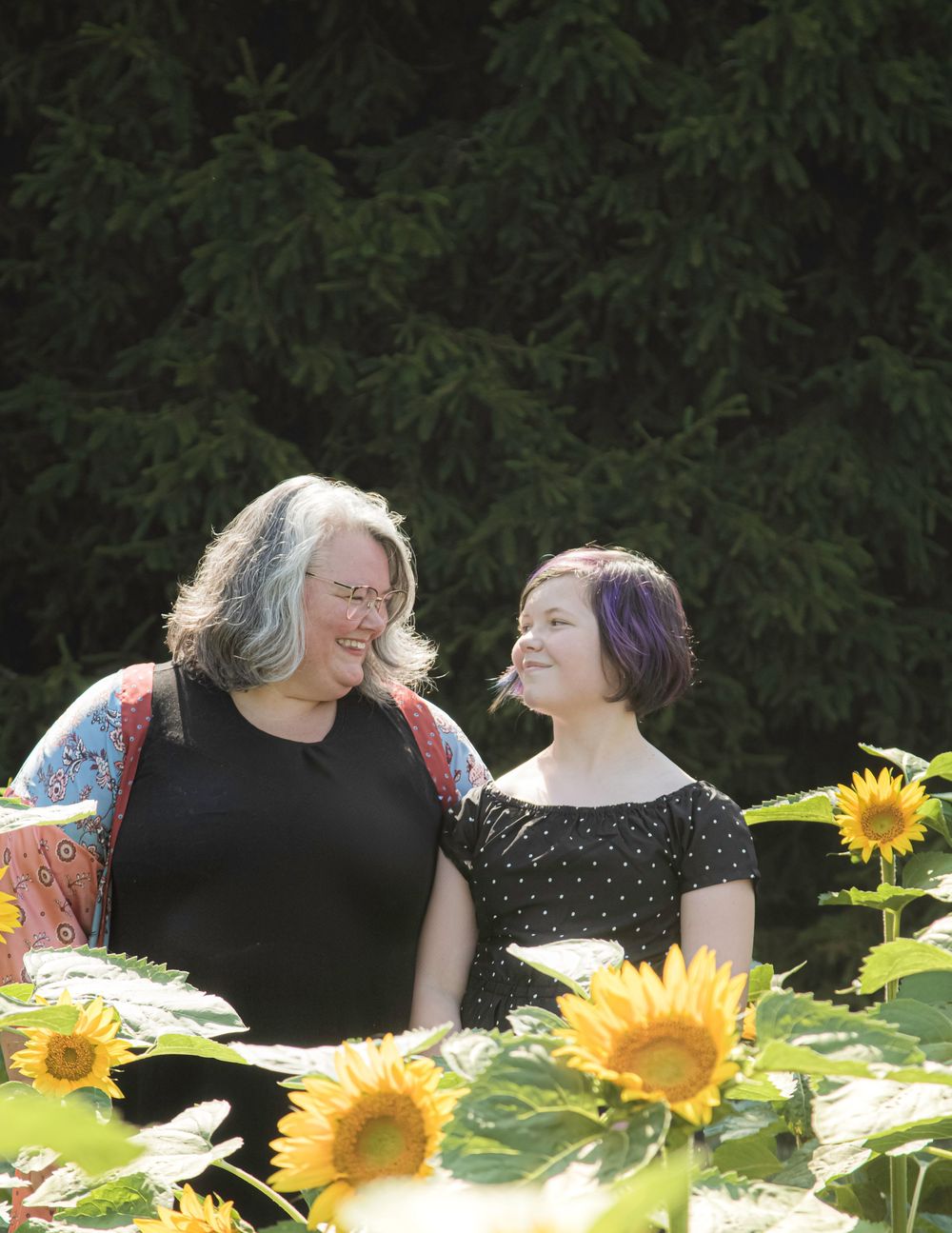 Two people share a joyful moment together among bright yellow sunflowers in a garden with evergreen trees in background.