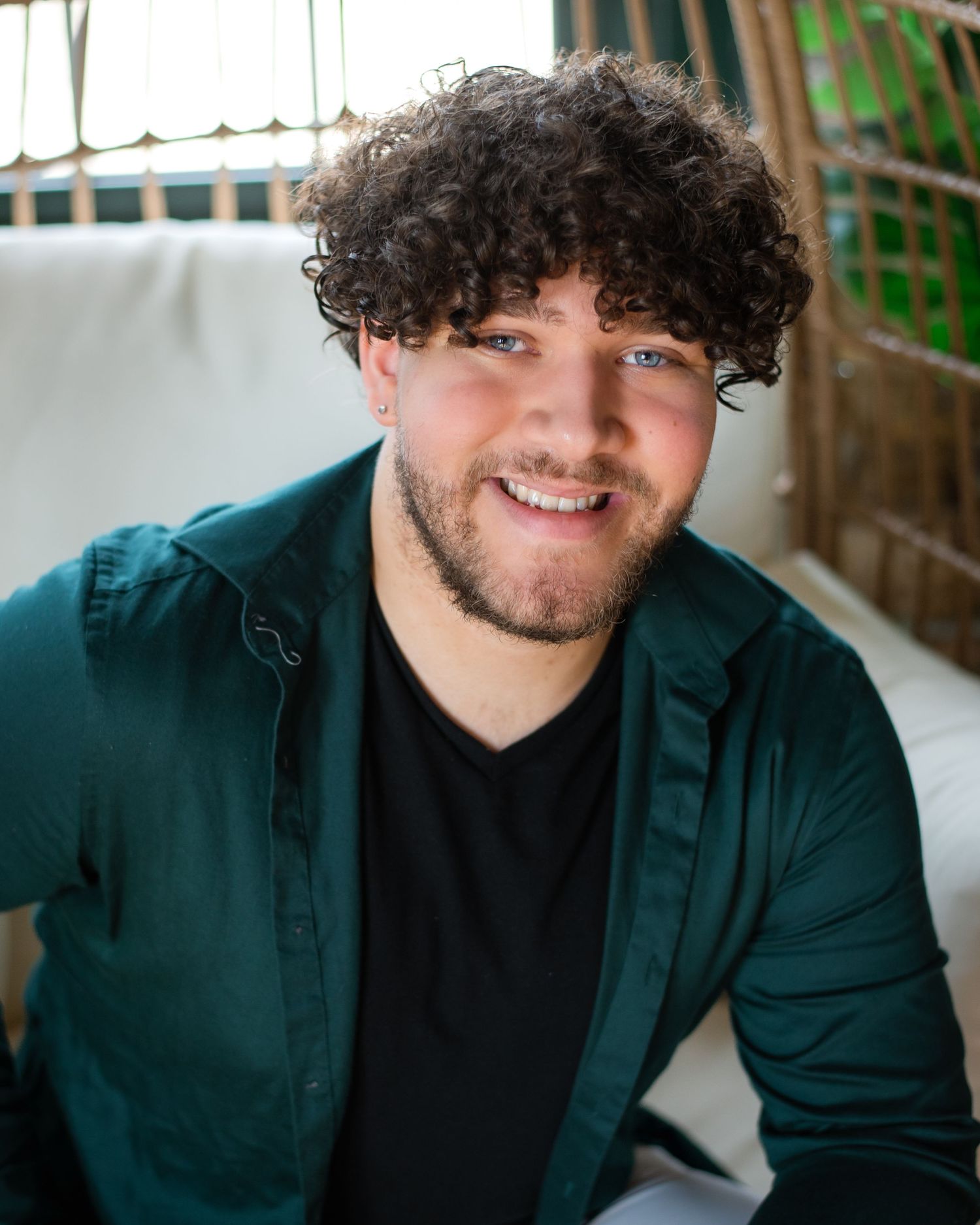 A person wearing a green blazer and black shirt smiles warmly in an indoor portrait setting.