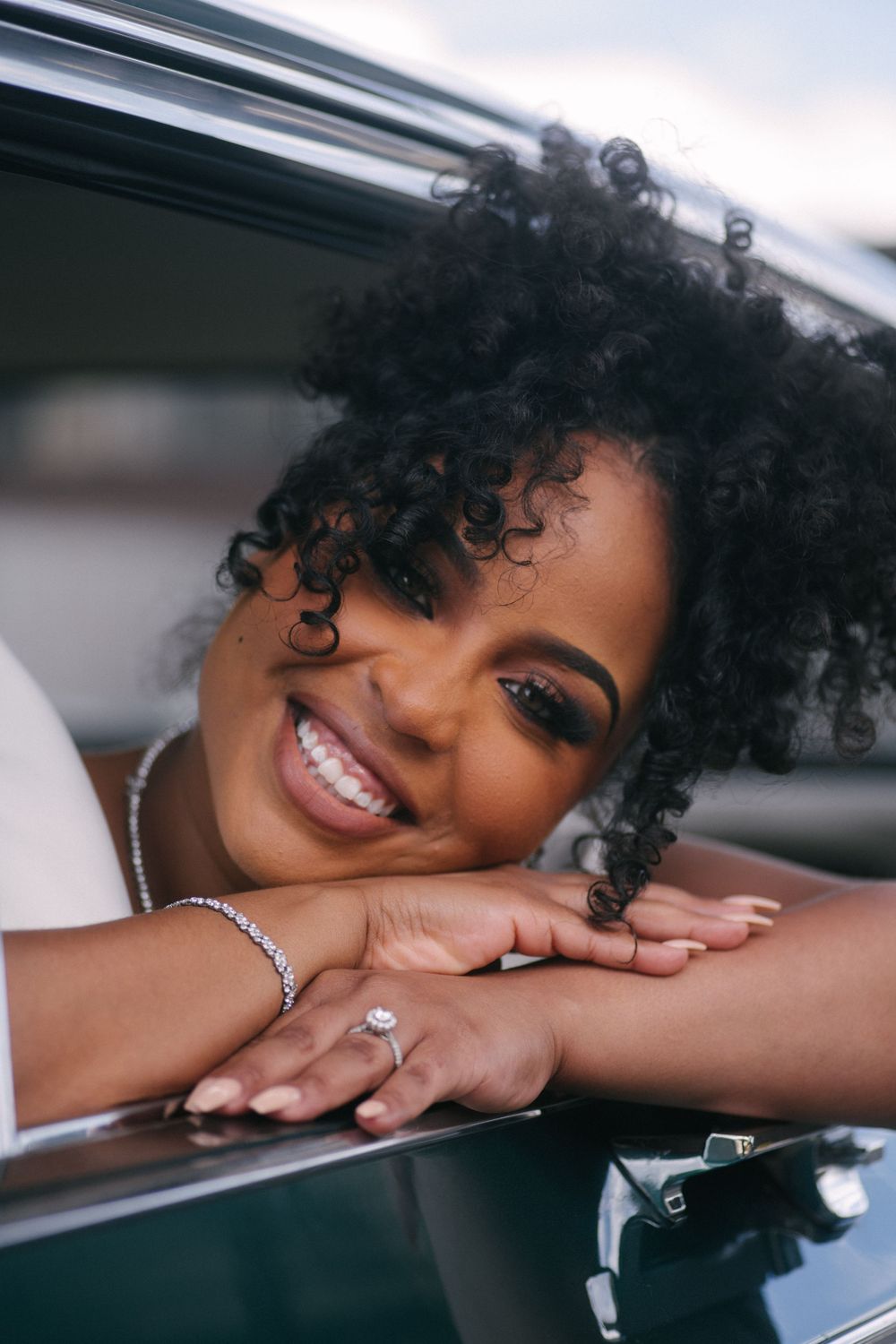 A smiling individual with natural curly hair leans on a car window while wearing a bracelet and ring.