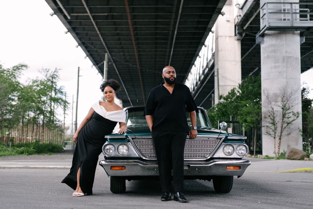 Stylish couple in black and white outfits pose together in front of a vintage car under an overpass.