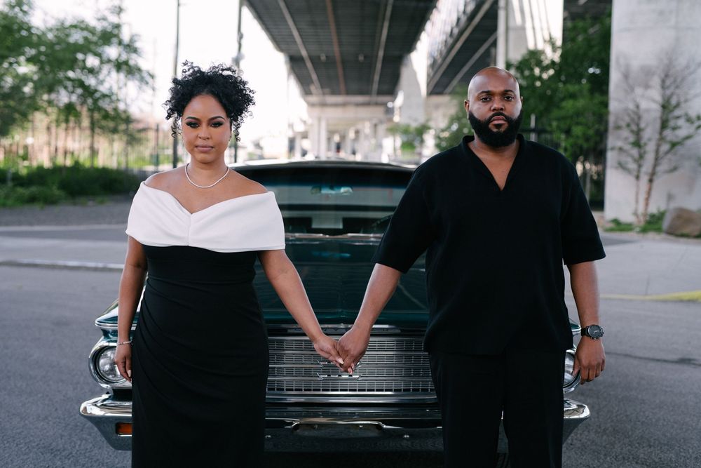 Couple in matching black attire hold hands while standing in front of a classic car beneath a bridge.