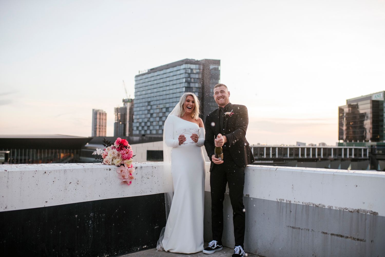 Wedding couple stands silhouetted against bright sky on modern rooftop venue with city skyline in background.