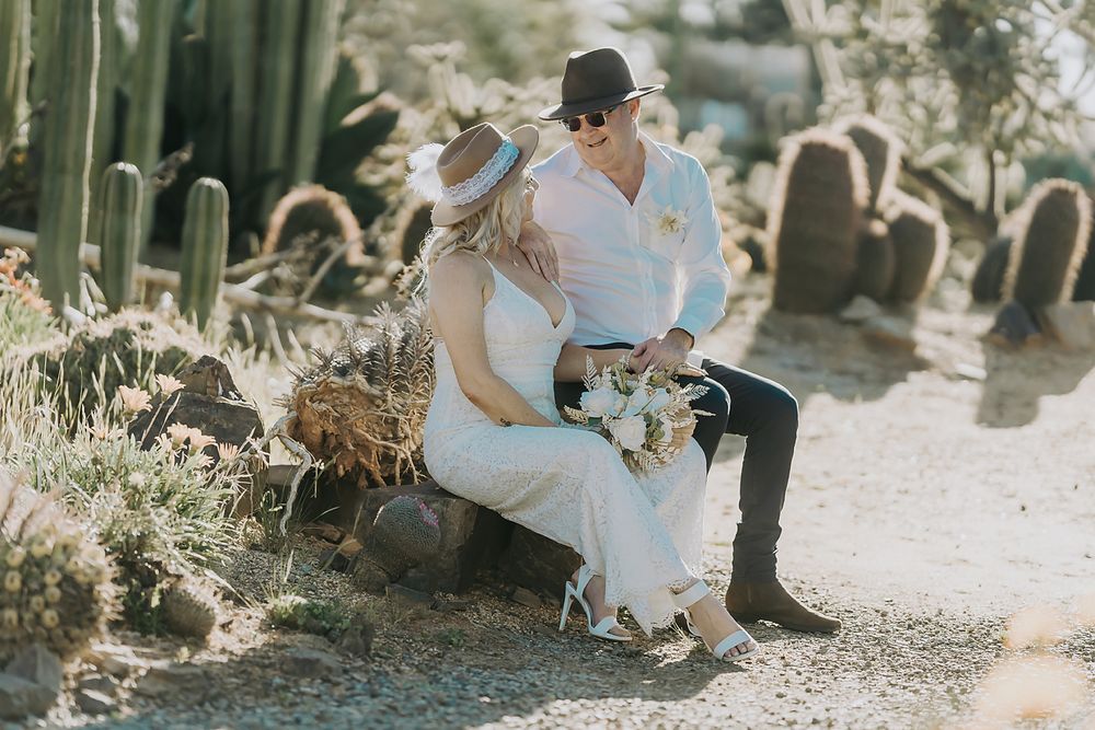 Romantic couple in western attire sitting together amid desert cacti at golden hour.