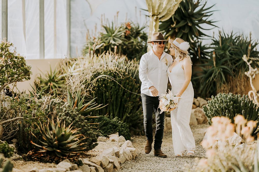 Wedding couple standing together in a lush garden with tropical plants and white picket fence.