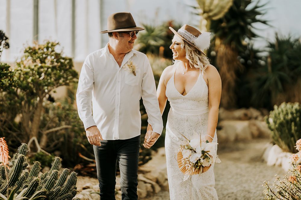 A couple walks hand in hand along a scenic desert path lined with tall cacti and tropical plants.