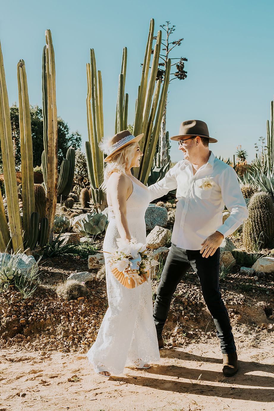 Wedding couple strolls through desert botanical garden with towering saguaro cactus in background.