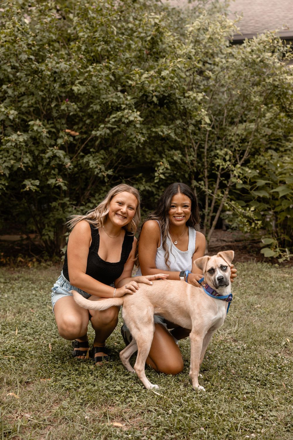 Two people in athletic wear pose outdoors on grass with a friendly tan-colored dog in a natural garden setting.