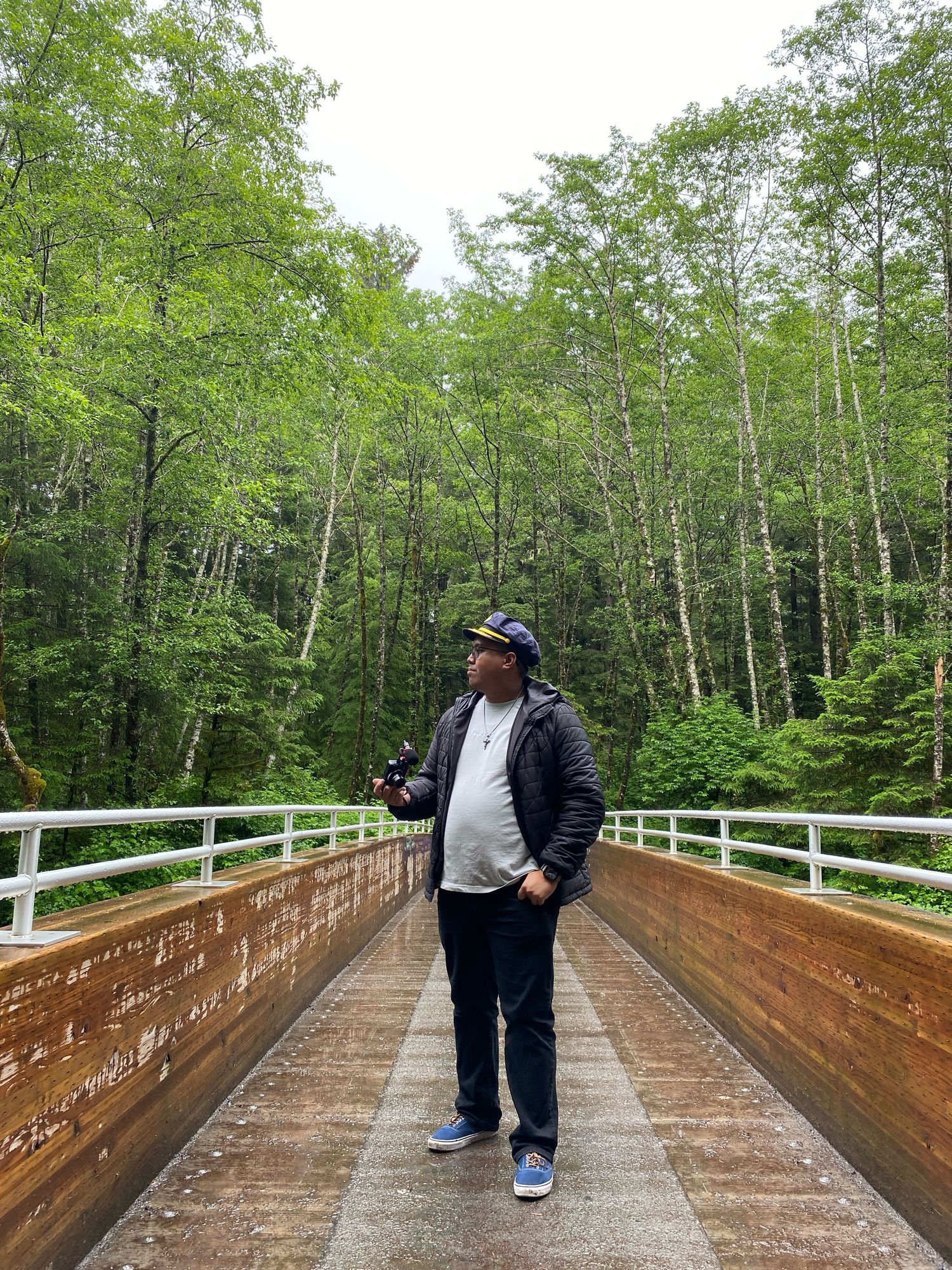 Person walking along a wooden boardwalk path through lush green forest wearing dark casual attire and sneakers.