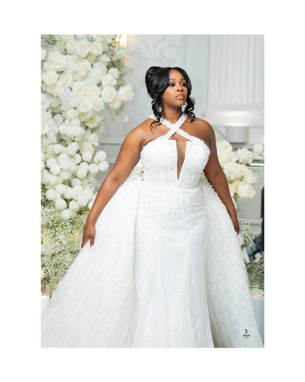A Black bride in a stunning white halter-neck beaded wedding gown with overskirt, posed before a floral backdrop.