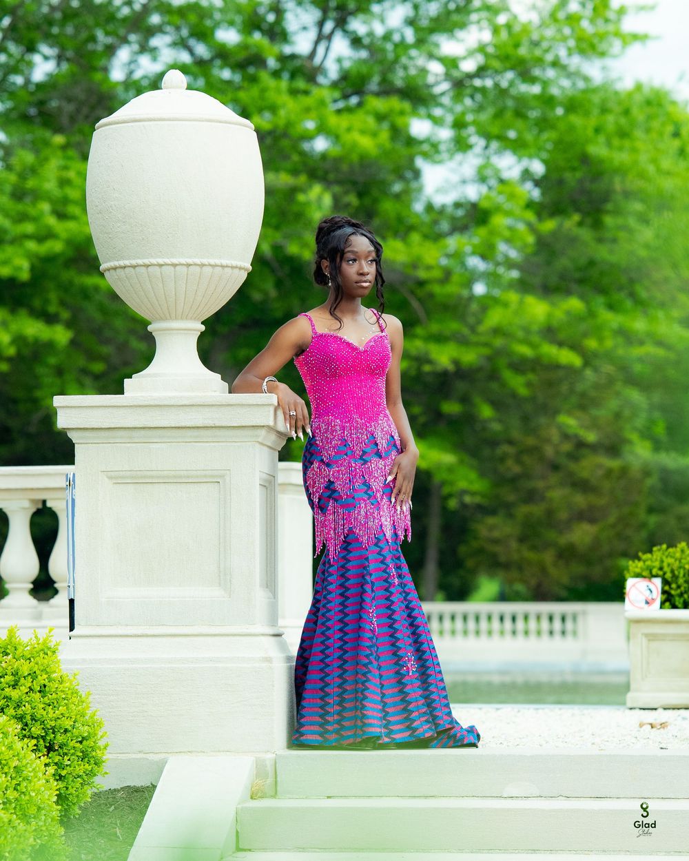 Woman in vibrant pink and purple African print mermaid gown posing elegantly by white stone pillar in lush garden.