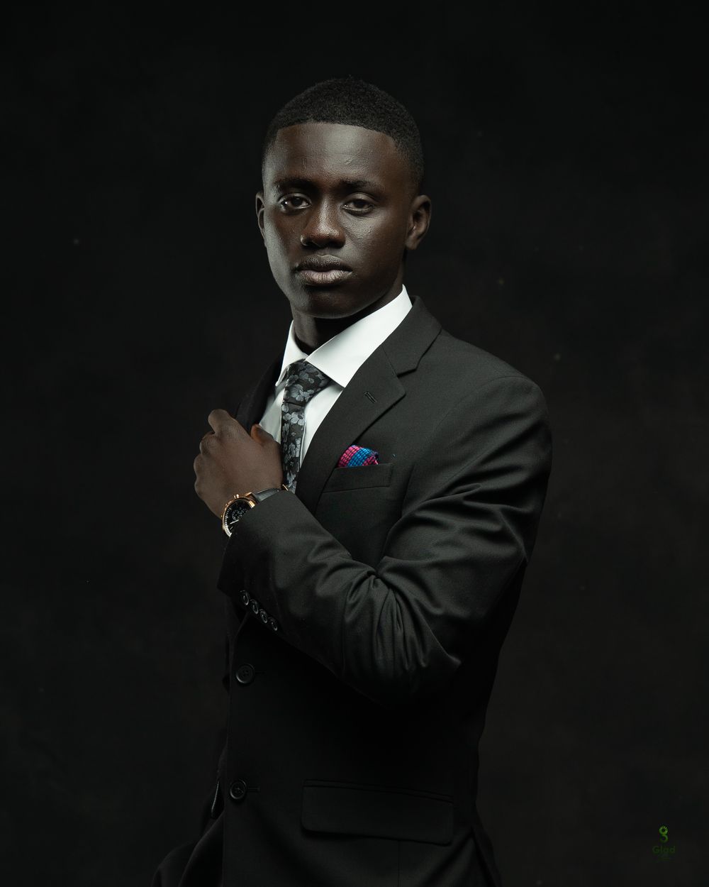 Young man in dark suit with patterned tie and blue pocket square, posing confidently against dark background.