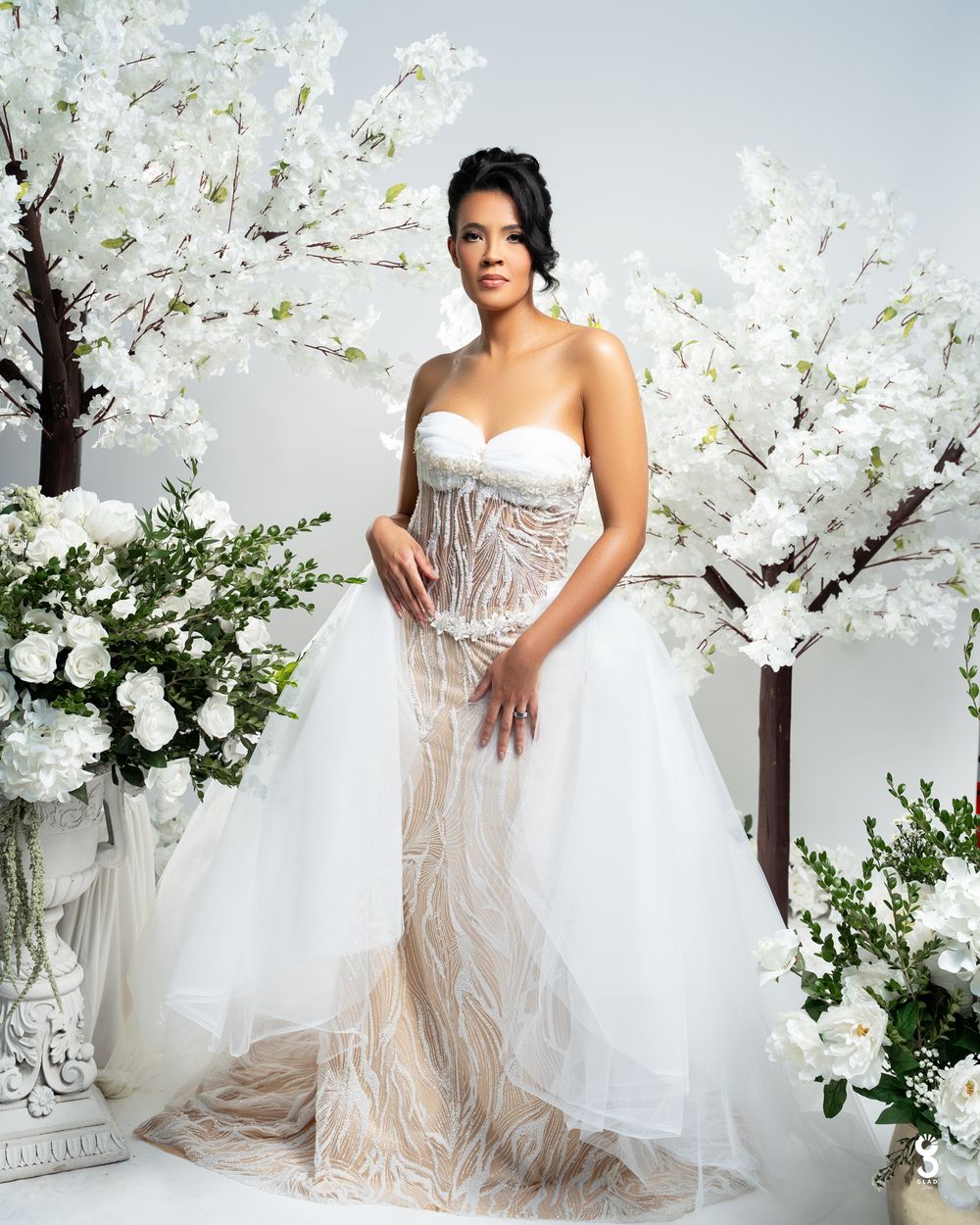 Bride in strapless beaded tulle wedding gown poses among white floral trees and roses in elegant bridal portrait.