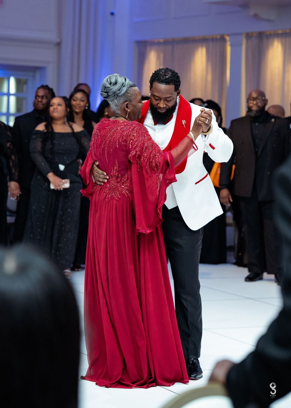A groom in a white tuxedo shares a mother-son dance with an elegant woman in a red gown at a wedding reception.