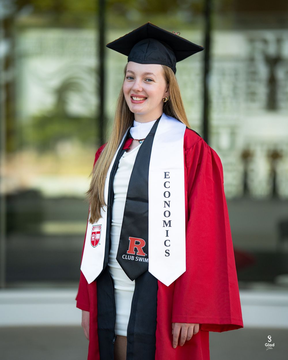 Rutgers University graduate in red cap and gown with Economics stole smiling at outdoor graduation photo.
