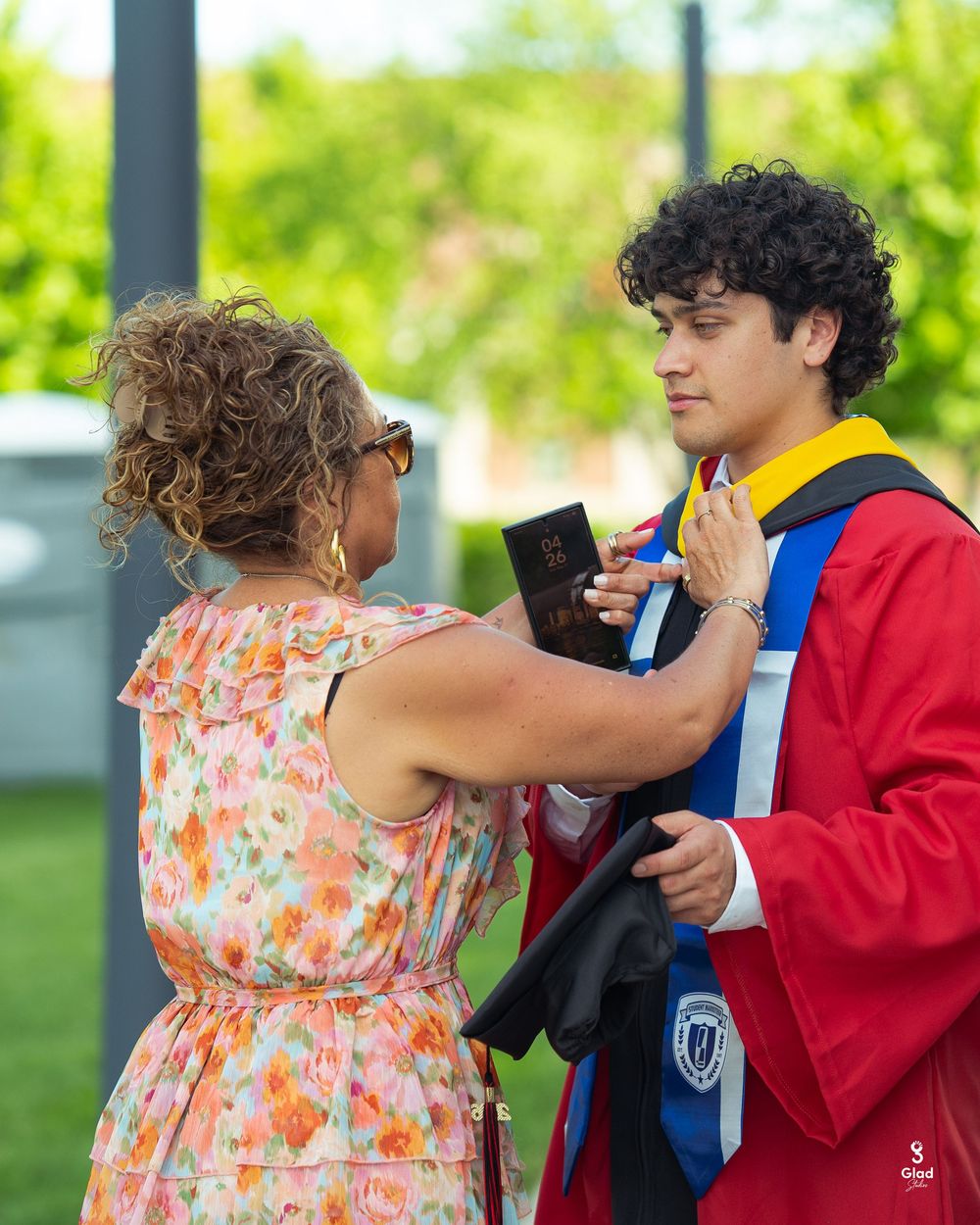 A proud mother adjusts her son's graduation regalia as he stands in a red cap and gown outdoors.