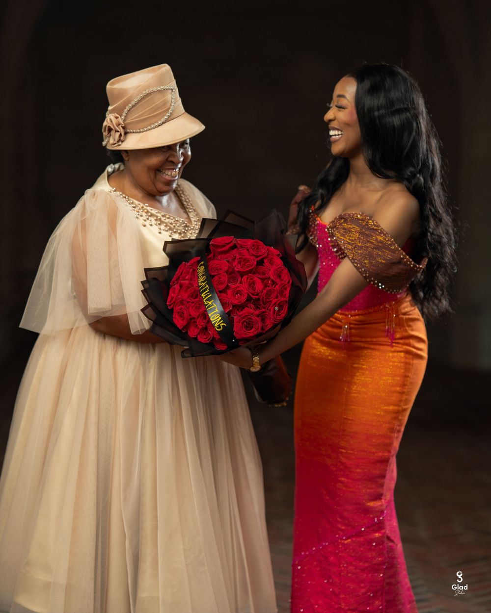 Two elegantly dressed women share a joyful moment as one presents a bouquet of red roses at a formal event.