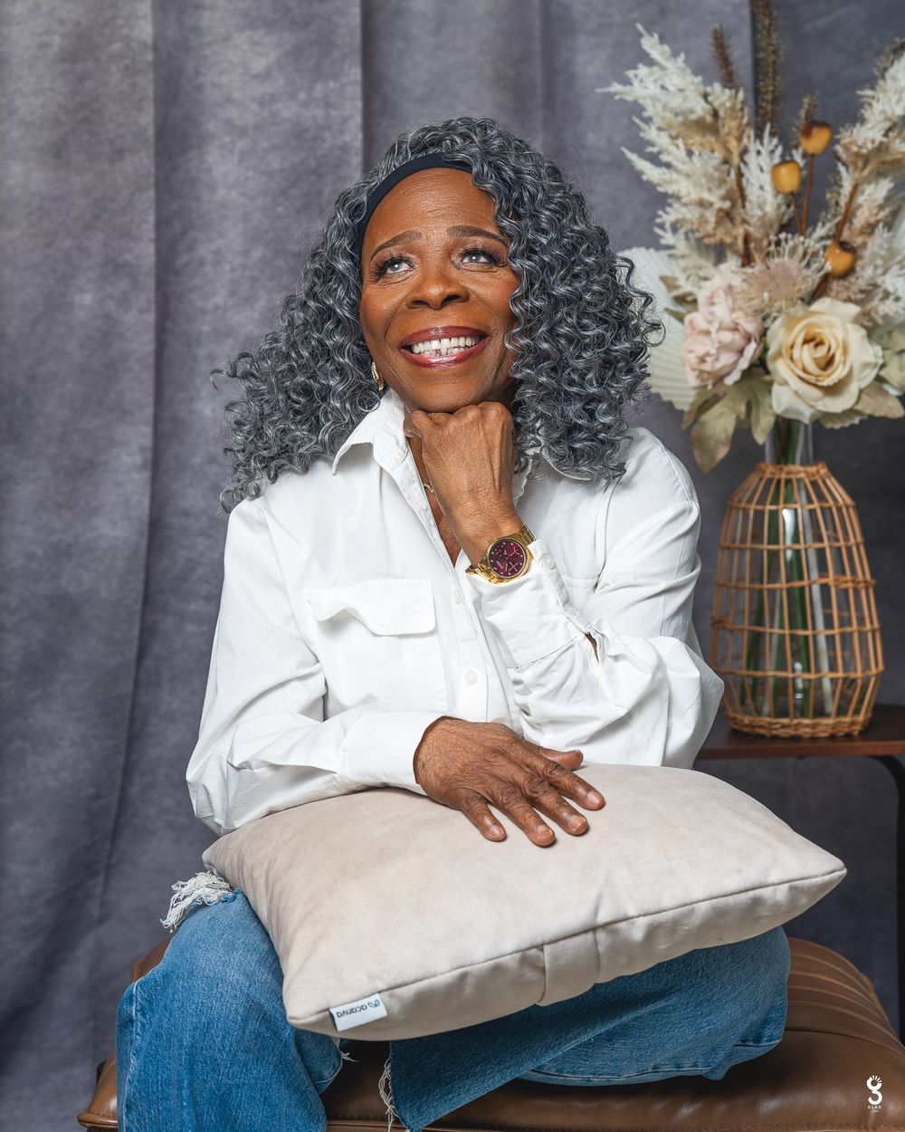 Smiling woman with gray curly hair holds a beige pillow, wearing white shirt and jeans, seated against gray backdrop.