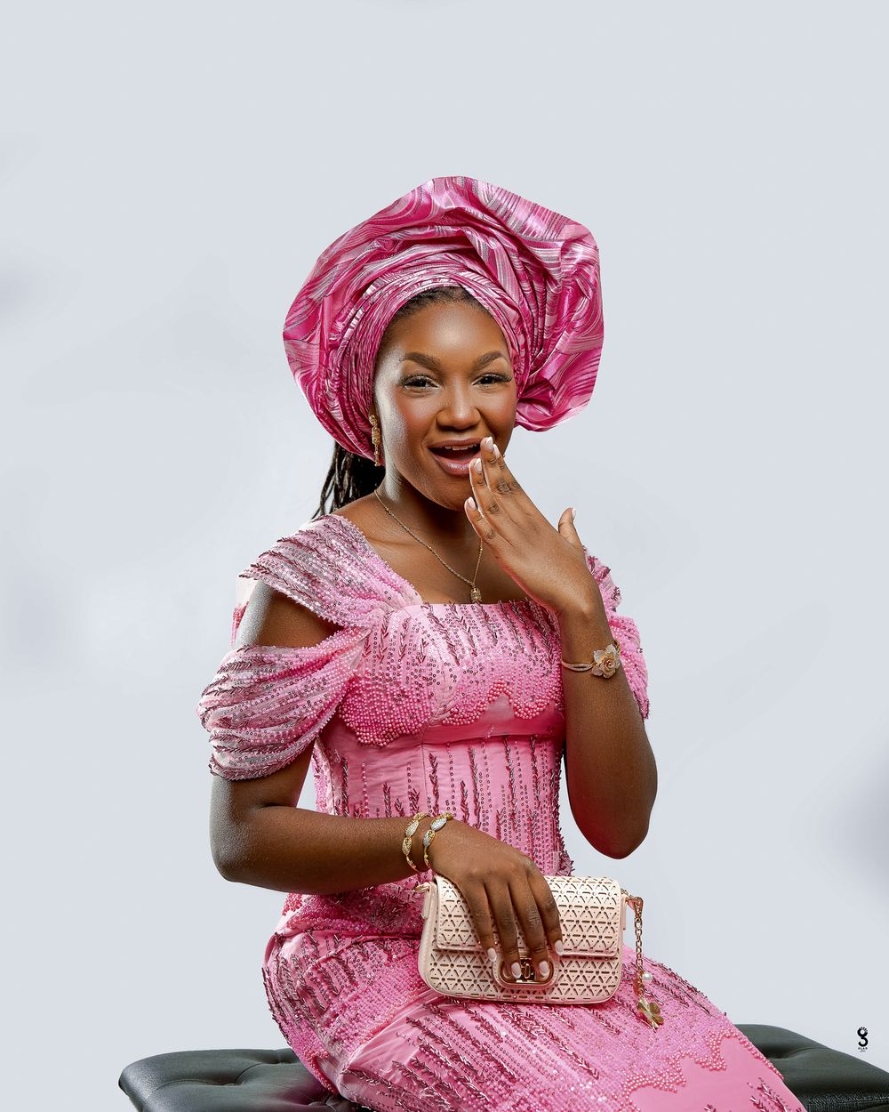 A woman in a pink lace dress and matching gele headwrap smiles joyfully, holding a beige handbag.