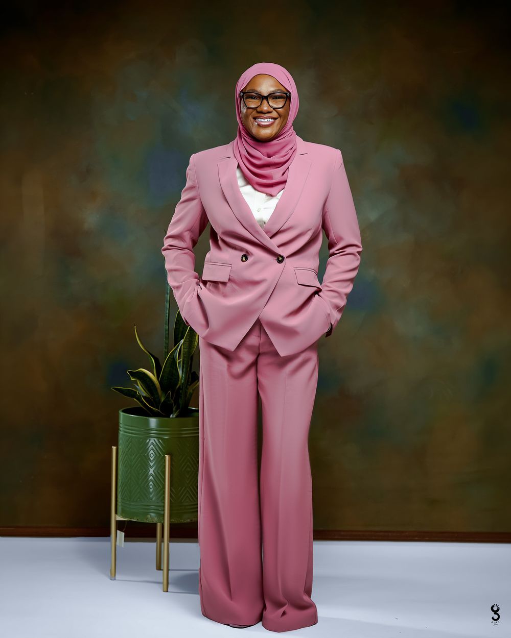 Woman in pink hijab and matching blazer pantsuit smiling confidently, posing beside a green plant in a studio.